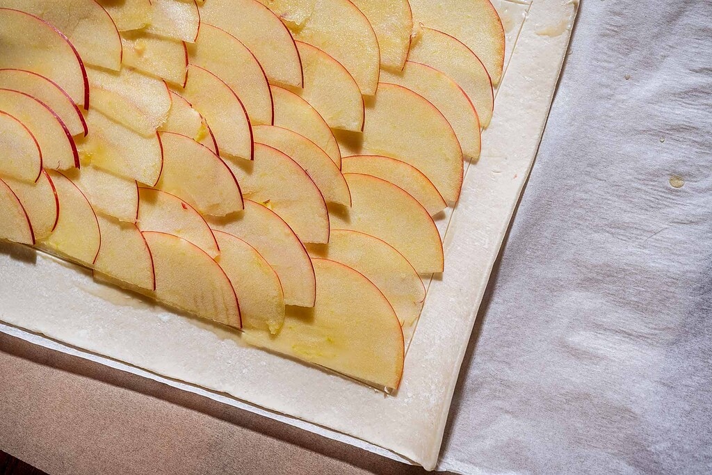 Thin apple slices arranged in overlapping rows on a sheet of pastry dough, placed on parchment paper and ready for baking—a delicious caramel apple tart in the making.