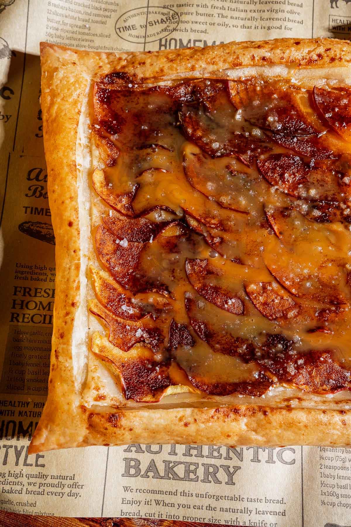 A rectangular caramel apple tart made with puff pastry, topped with caramelized apple slices and a glossy glaze, rests on printed parchment paper adorned with bakery-themed text.