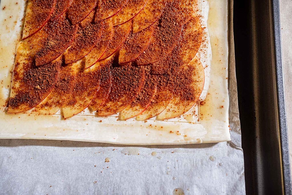 A sheet of puff pastry topped with thinly sliced apples and a layer of cinnamon sugar, arranged on a baking sheet lined with parchment paper, creates the base for a delicious caramel apple tart.