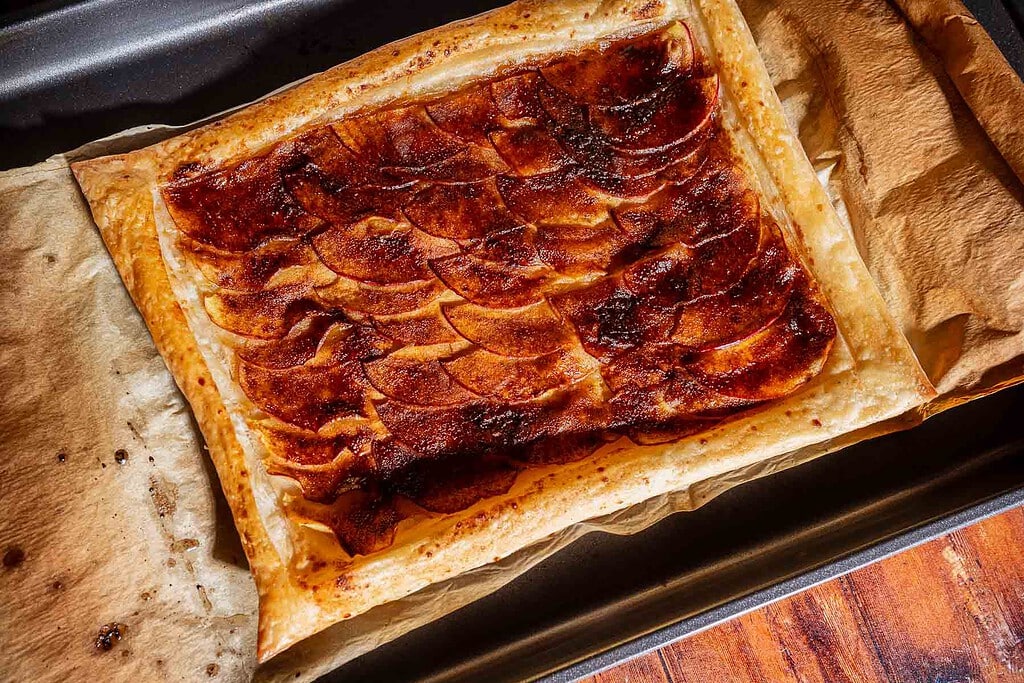 Rectangular caramel apple tart with thinly sliced apples on puff pastry, baked to golden perfection and placed on parchment paper in a baking tray.