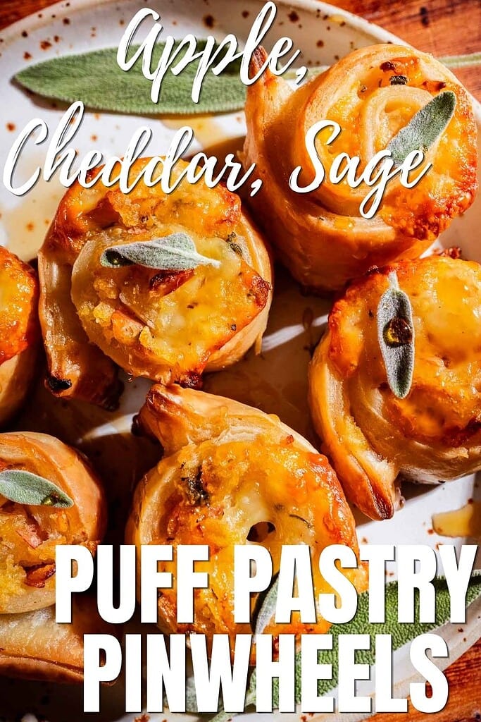 A plate of golden, flaky puff pastry pinwheels filled with apple, cheddar, and sage, each beautifully topped with a fresh sage leaf. Text on the image reads: “Apple, Cheddar, Sage Puff Pastry Pinwheels.”.