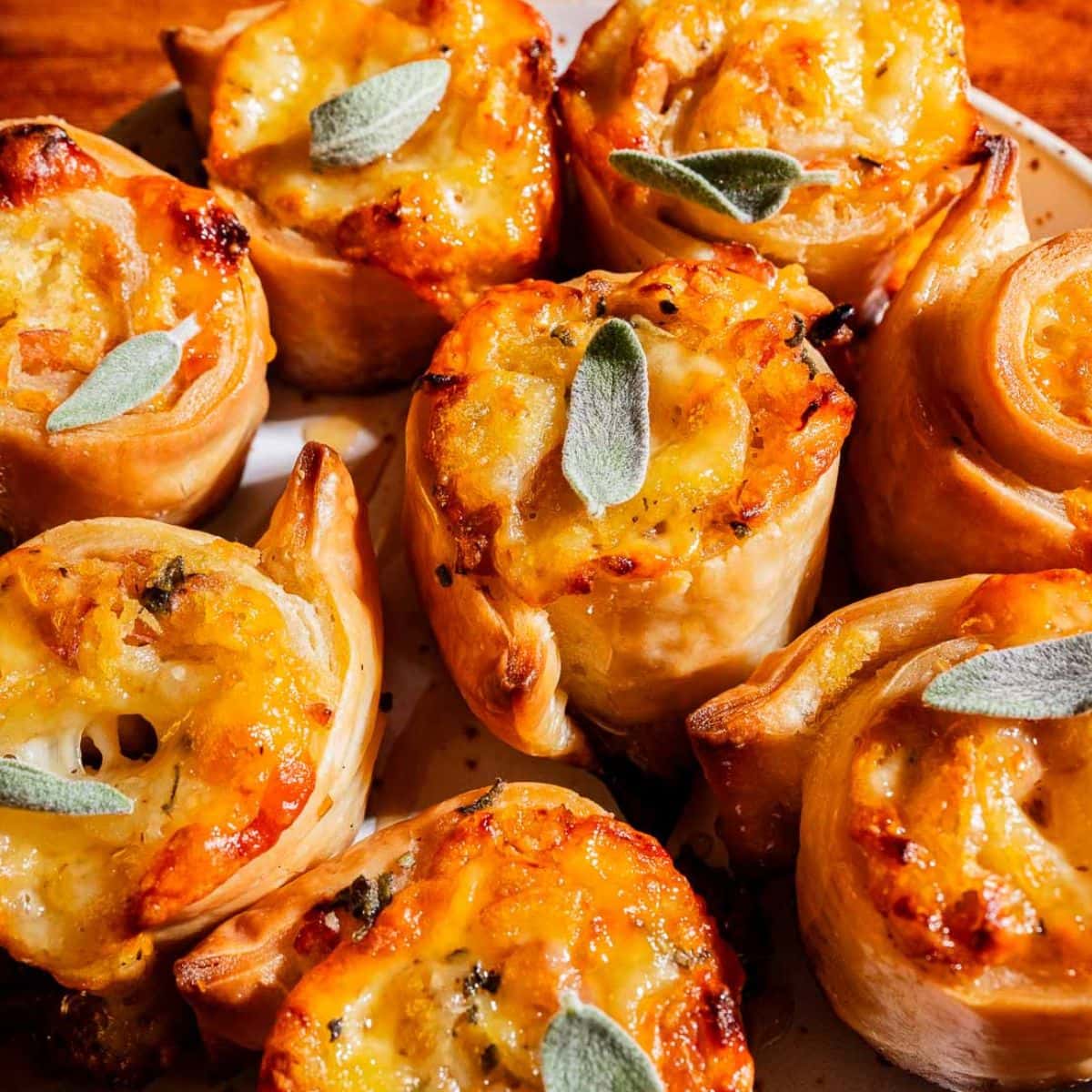 A close-up of several golden-brown puff pastry pinwheels topped with melted cheese and small sage leaves on a white plate.