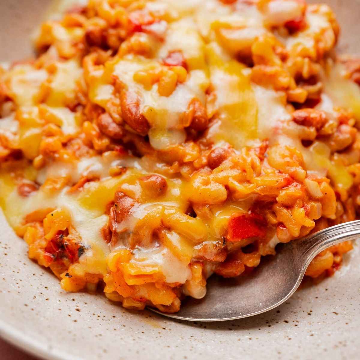 A close-up of cheesy Mexican rice with beans and vegetables, reminiscent of slow cooker chili mac, topped with melted cheese and served on a speckled plate with a spoon.