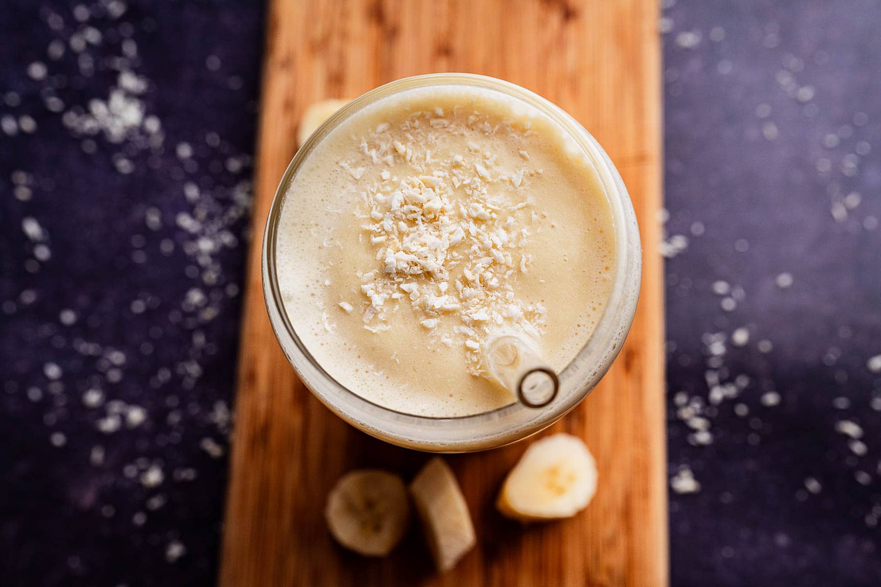 A glass of tropical smoothie, brimming with banana flavors and topped with shredded coconut, is placed on a wooden board with banana slices nearby.