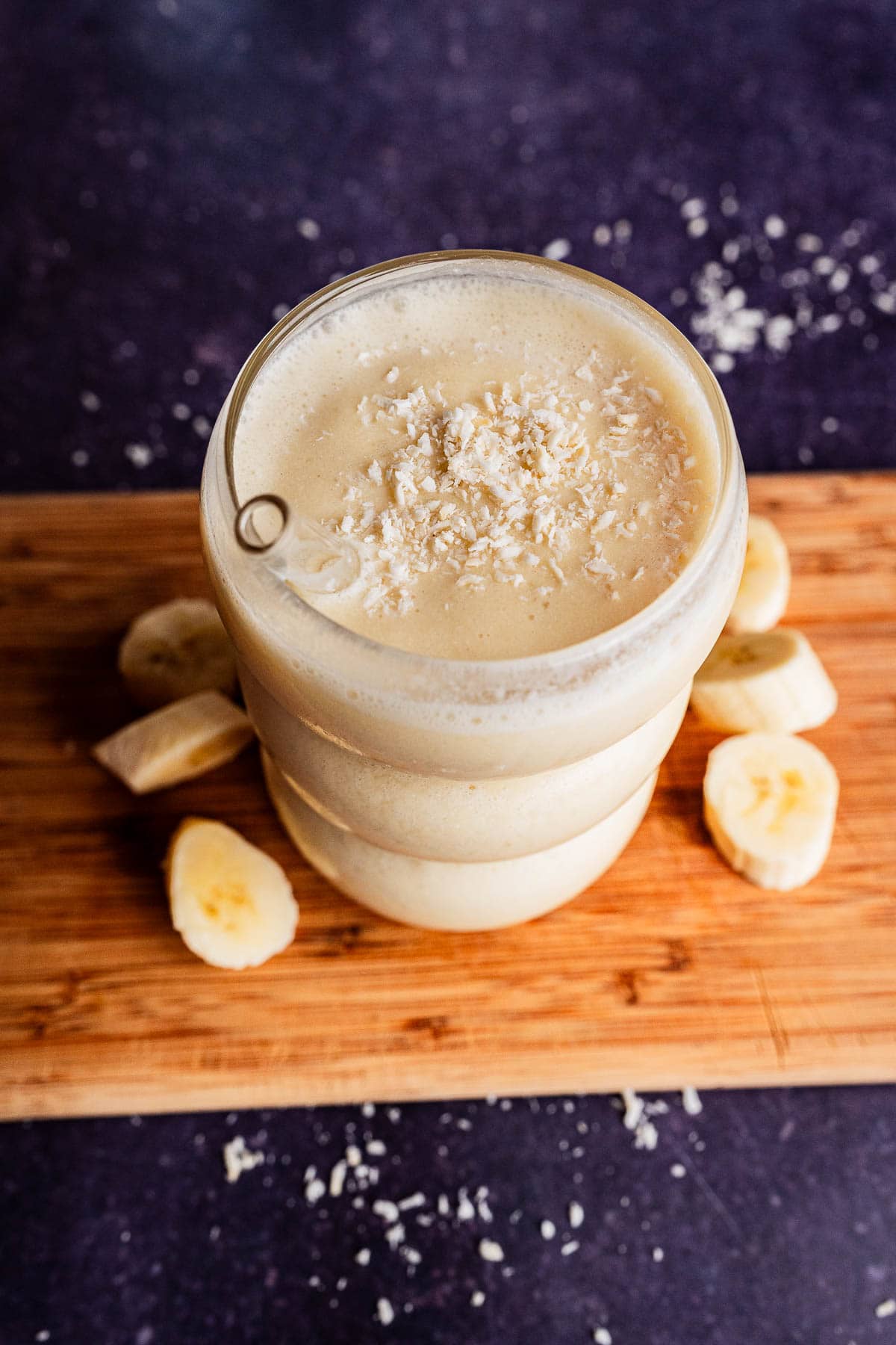 A glass of tropical smoothie topped with shredded coconut, placed on a wooden board with banana slices around it, against a dark background.