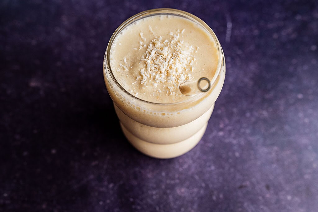 A glass filled with a creamy light brown tropical smoothie topped with shredded coconut, placed on a dark textured surface.