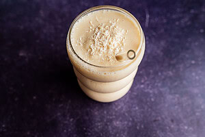 A glass filled with a creamy light brown tropical smoothie topped with shredded coconut, placed on a dark textured surface.