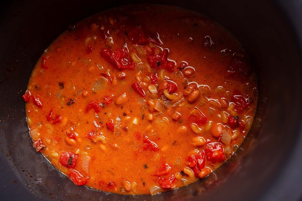 A close-up of bean and tomato stew with a red, soupy base, visible diced tomatoes, beans, and onions simmering inside a dark pot, reminiscent of a hearty slow cooker chili mac.