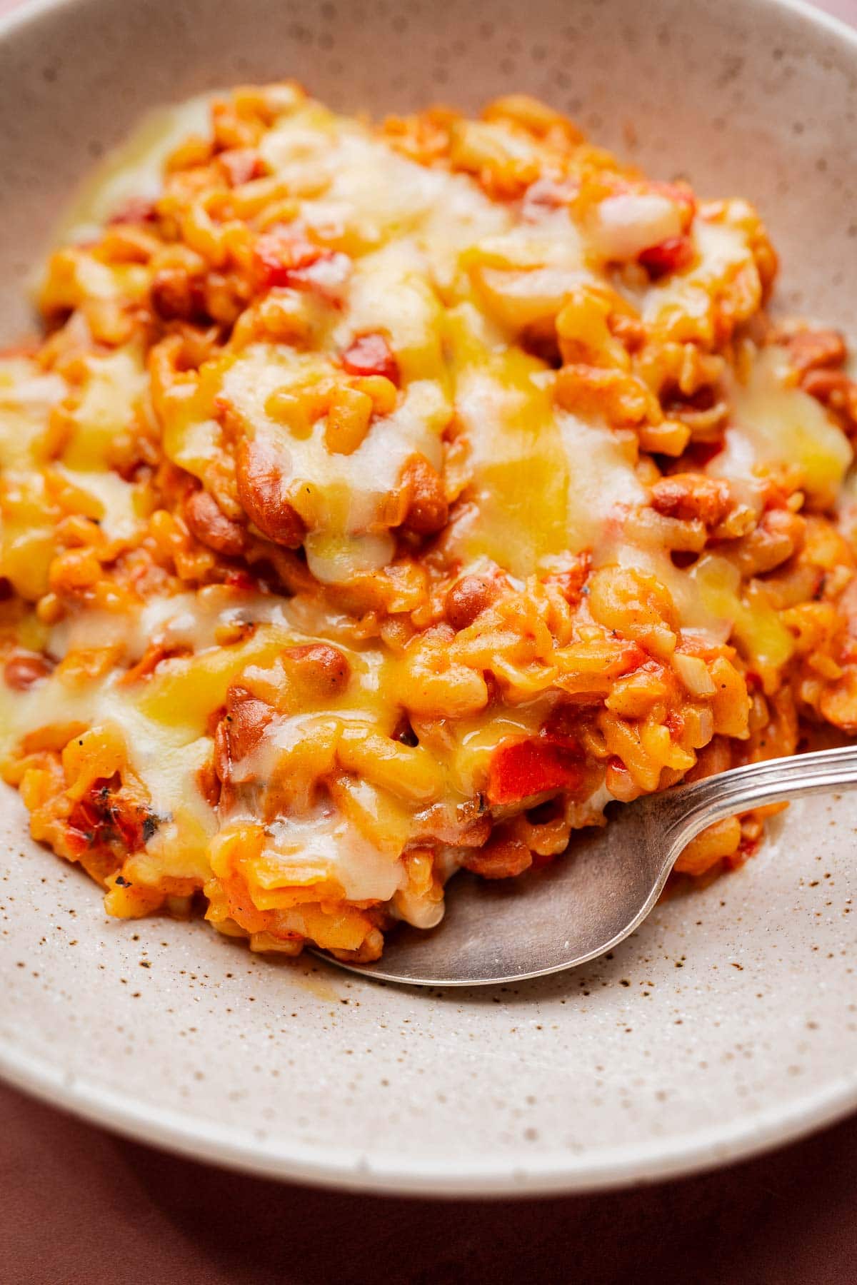 A close-up of a bowl of cheesy rice and beans with red bell peppers, topped with melted cheese, and a spoon resting in the food—reminiscent of a hearty slow cooker chili mac.