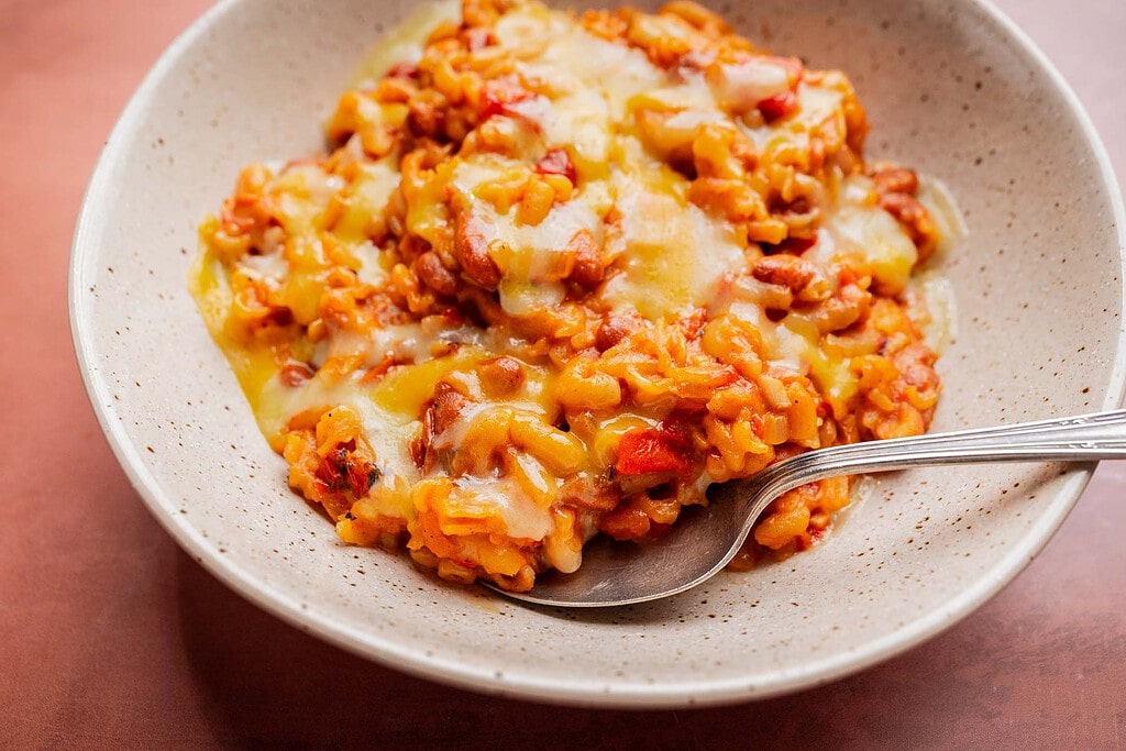 A bowl of cheesy macaroni with tomato sauce and ground meat, reminiscent of a comforting slow cooker chili mac, served with a spoon on a speckled ceramic plate.