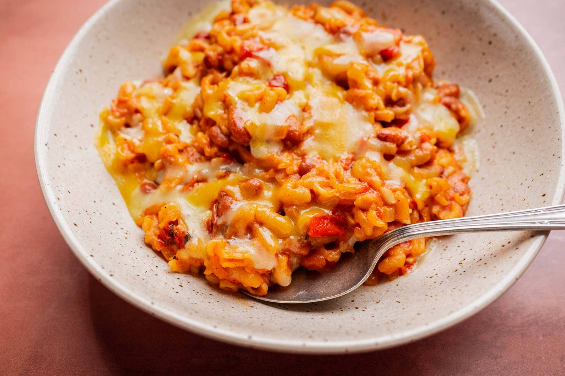 A bowl of cheesy macaroni with tomato sauce and ground meat, reminiscent of a comforting slow cooker chili mac, served with a spoon on a speckled ceramic plate.