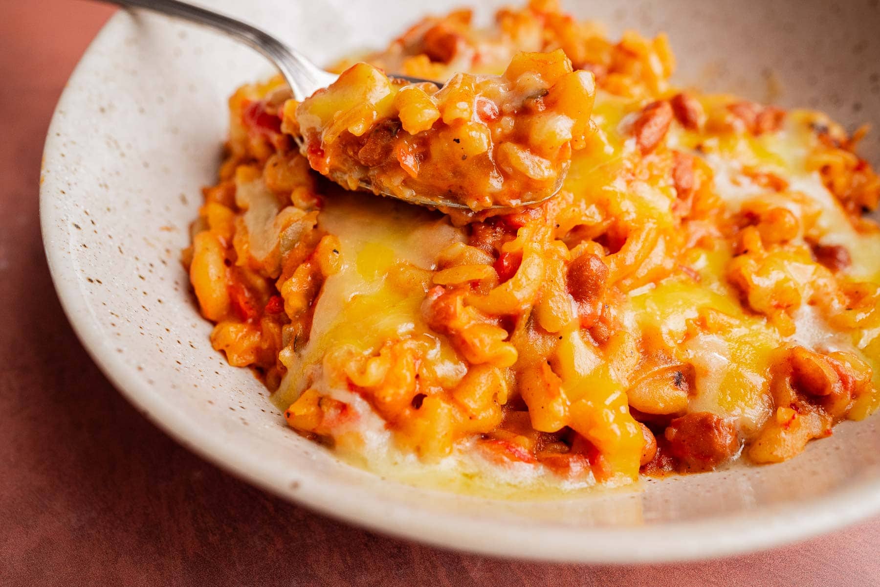 A close-up of baked cheesy pasta with beans and tomatoes in a ceramic bowl, reminiscent of slow cooker chili mac, with a spoon lifting a portion.