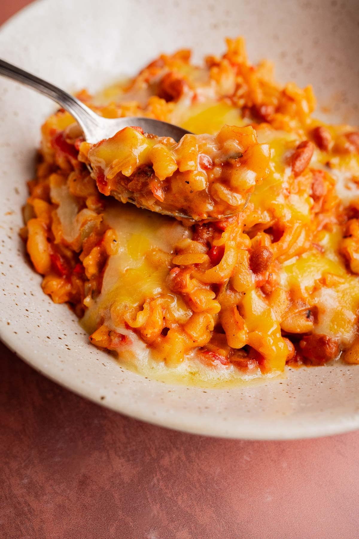A close-up of cheesy baked macaroni with tomato sauce and red beans on a speckled plate, reminiscent of hearty slow cooker chili mac, with a fork lifting a portion.