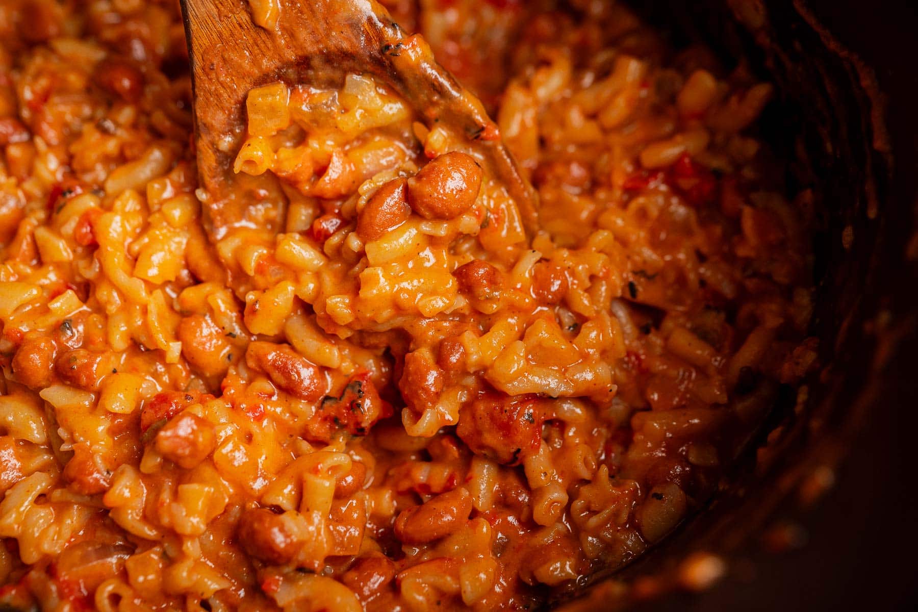 Close-up of a wooden spoon stirring a creamy mixture of rice, beans, and tomato sauce, reminiscent of a hearty slow cooker chili mac.