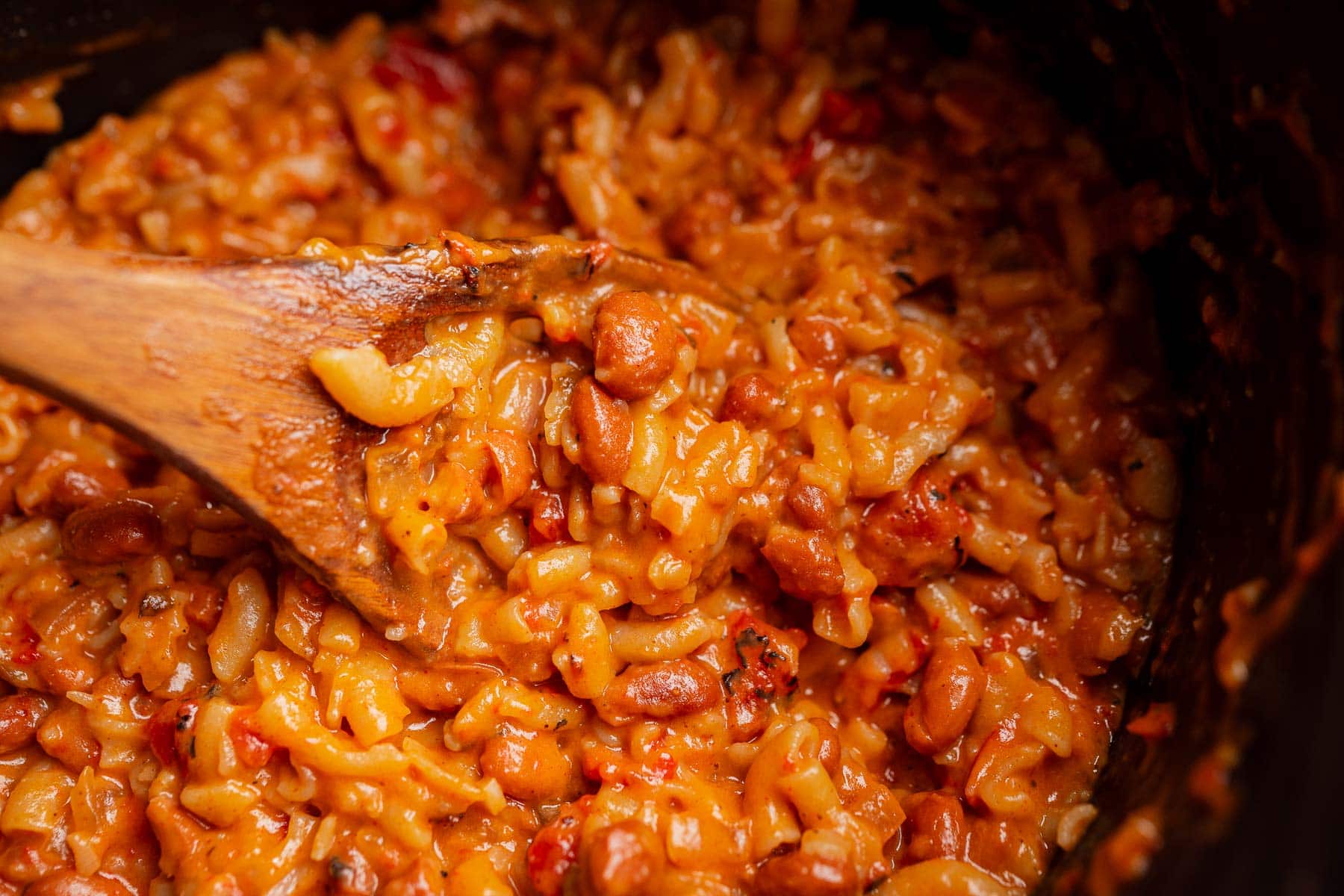 Close-up of creamy, cheesy rice and beans being stirred with a wooden spoon in a pot, reminiscent of a comforting slow cooker chili mac.