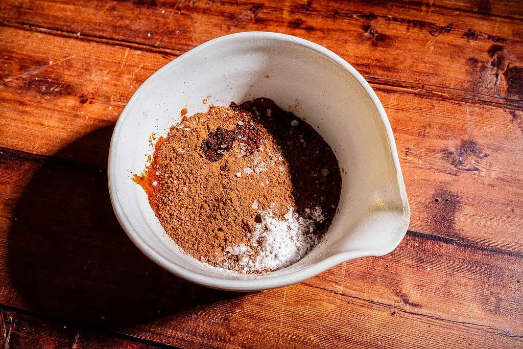 A white mixing bowl containing various dry baking ingredients, including cocoa powder, sugar, and baking powder, sits on a wooden surface.