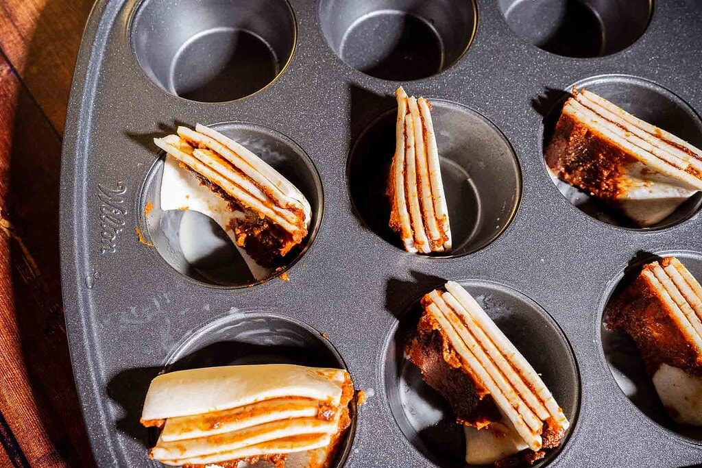 A muffin tin holds several stacks of alternately layered dough and filling, some placed in separate cups, in bright lighting.