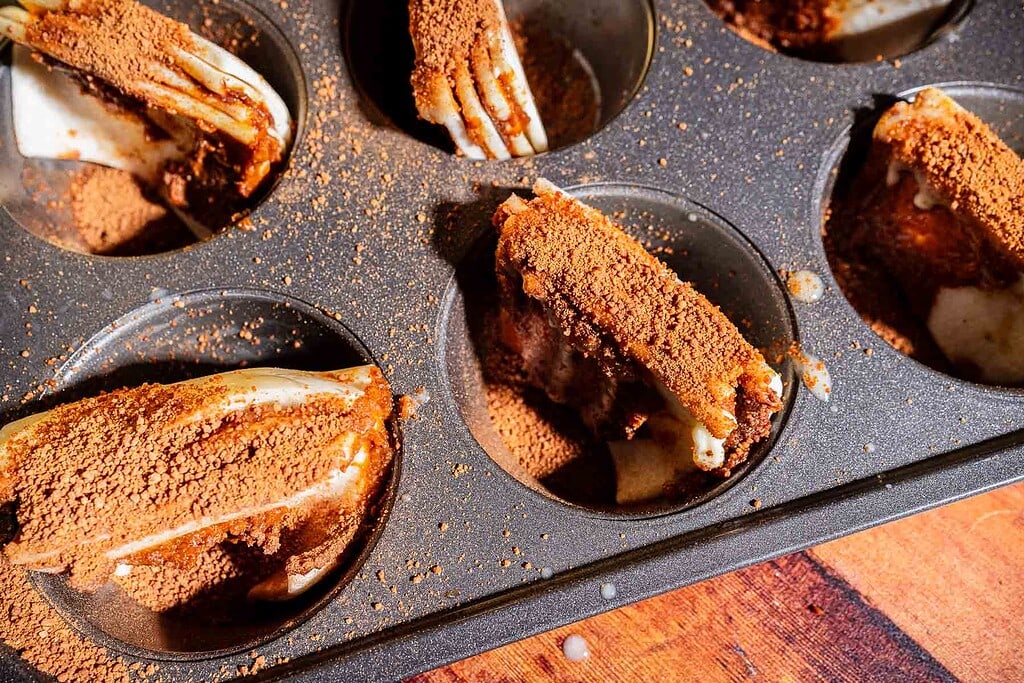 Close-up of cannoli shells filled with cream and topped with cocoa powder in a dark muffin tin on a wooden surface.