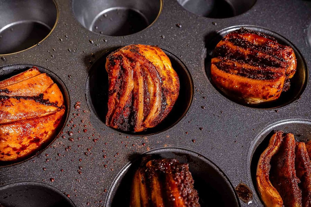 Slices of cooked bacon arranged in a muffin tin, each compartment holding a portion. The bacon appears crispy and seasoned, with some grease visible on the tray.