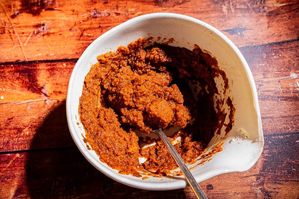 A white mixing bowl with a reddish-brown paste and a metal spoon, on a wooden surface.