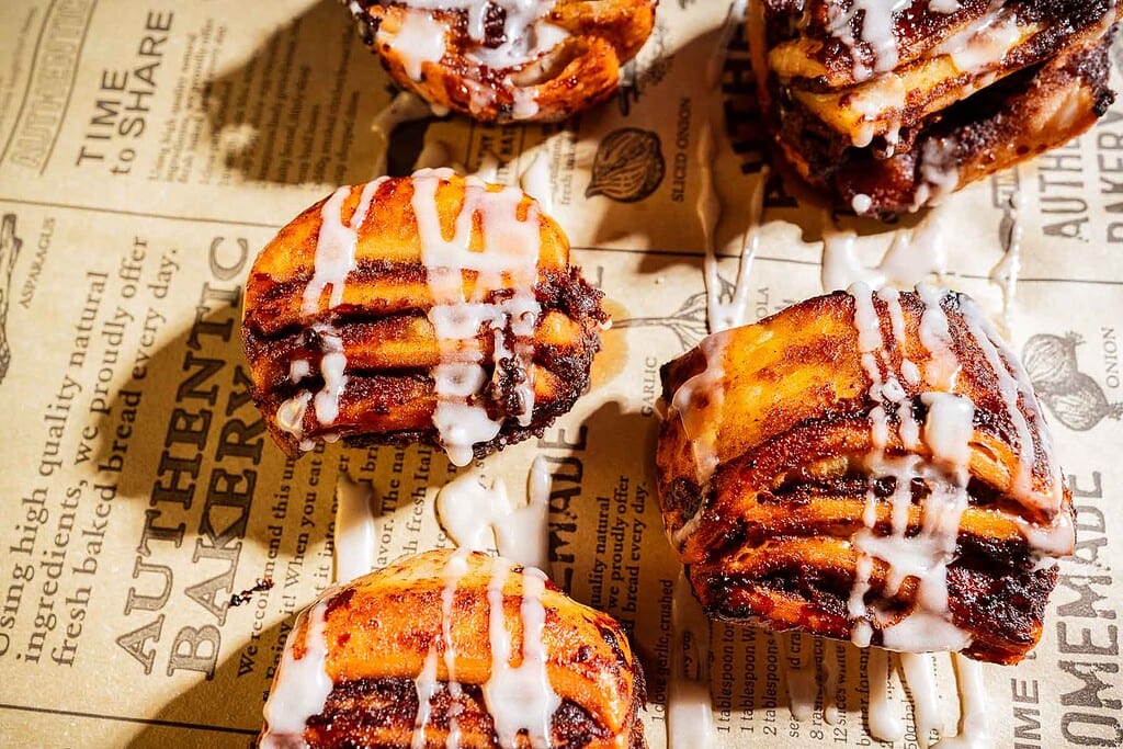 Four cinnamon rolls with icing are placed on a sheet of printed parchment paper, viewed from above in bright lighting.