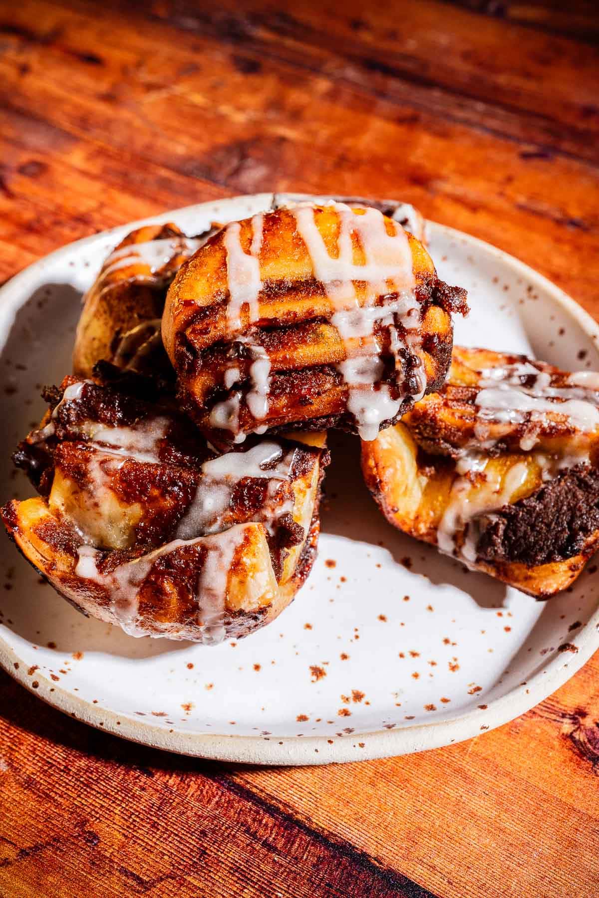 Four puff pastry pull aparts with icing are stacked on a white speckled plate, set on a wooden surface.