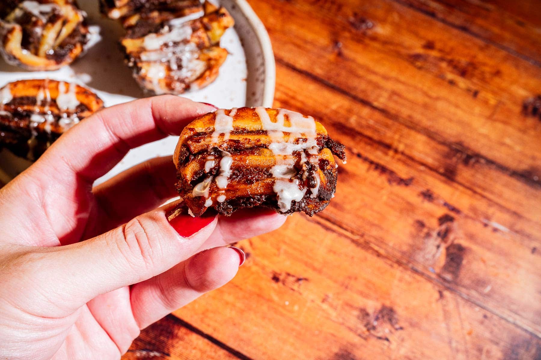 A hand holding a small pumpkin pastry with icing, with more rolls on a plate in the background on a wooden table.