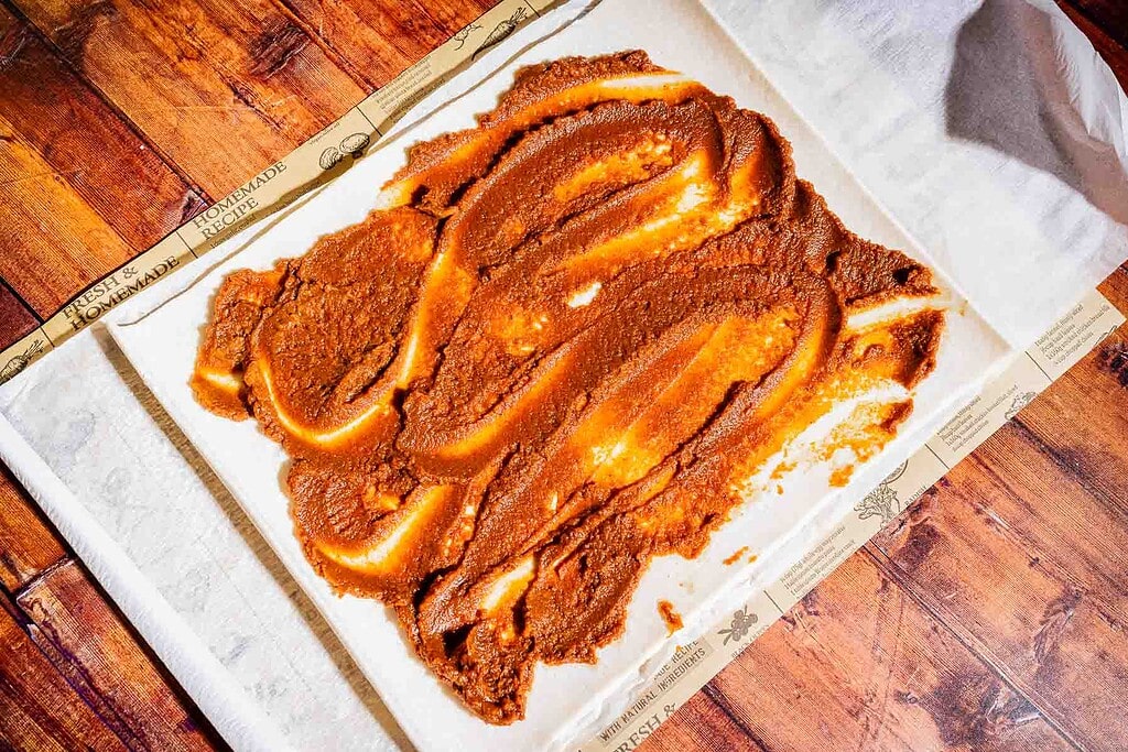 A thick layer of reddish-brown paste is spread unevenly on parchment paper placed on a wooden surface.