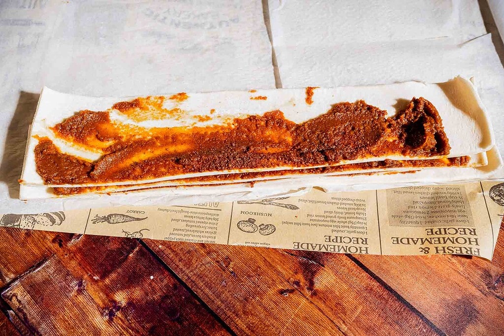 Sheet of pastry dough partially rolled with a layer of reddish-brown filling, sitting on a sheet of parchment paper on a wooden surface.