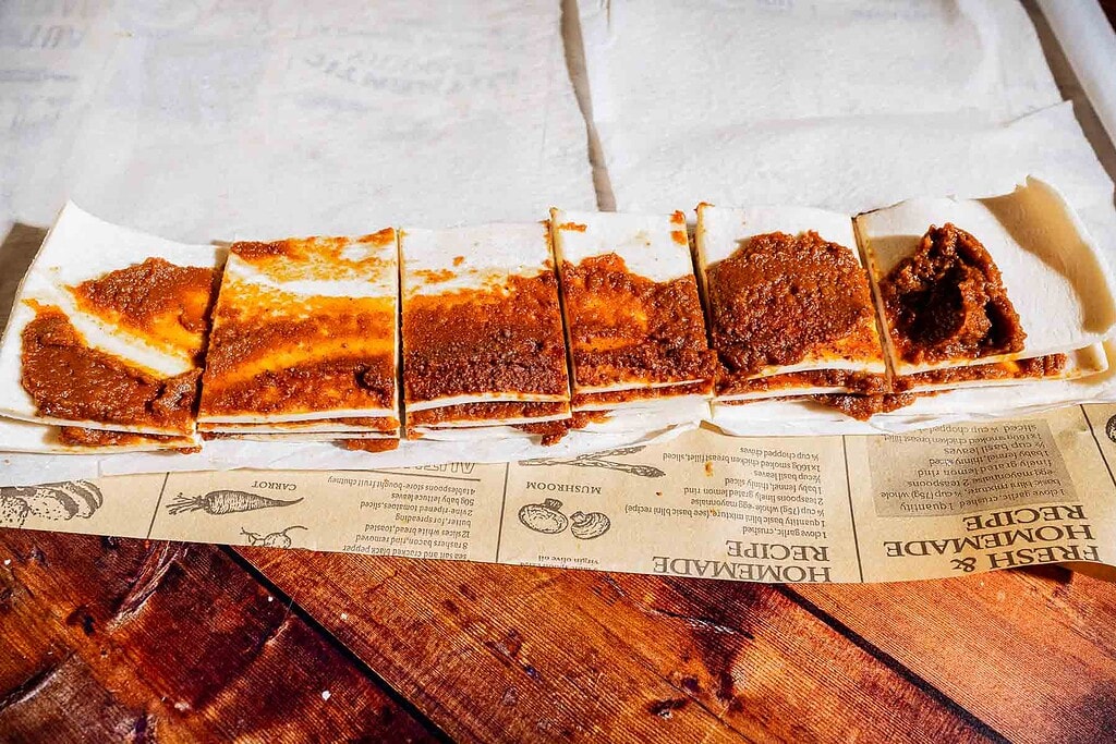 Several rectangular pieces of flatbread with a red-brown spread are arranged in a row on parchment paper atop a wooden surface.
