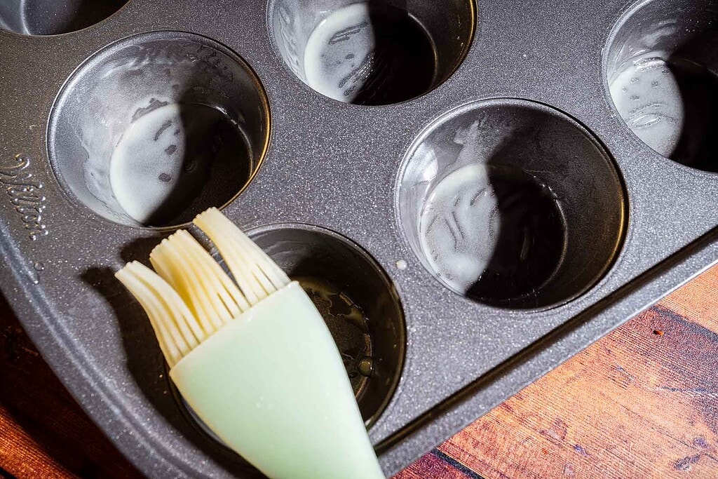 A muffin tin with several cups brushed with oil or butter, a silicone pastry brush resting in one of the cups, on a wooden surface.
