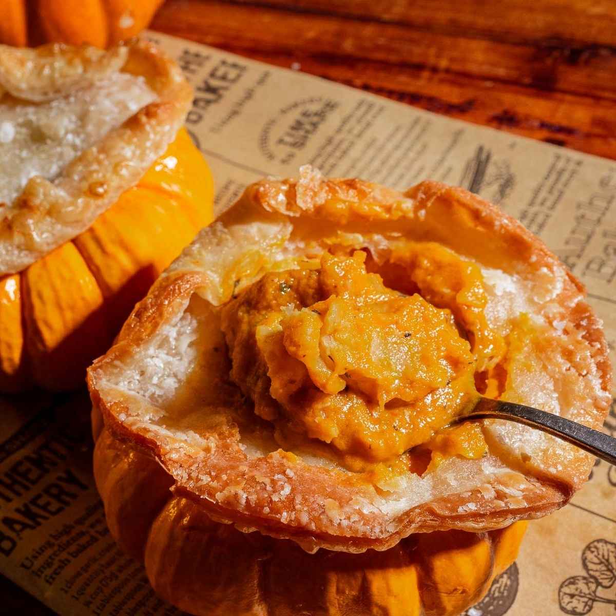 A spoon scoops pumpkin soup from a baked pumpkin with a golden puff pastry top, resting on parchment paper with printed text.