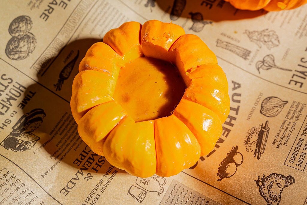 A round pumpkin with the top cut off and filled with a thick pumpkin soup sits on printed brown paper featuring text and food illustrations, making it a delightful example of mini pumpkin bowls.