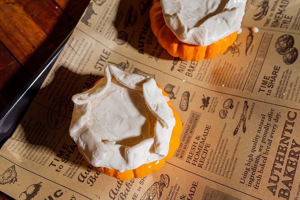 Two small pumpkins with cream-colored pastry dough draped over the tops, resembling puff pastry tops, are placed on a vintage-style printed baking sheet—perfect for serving pumpkin soup in charming mini pumpkin bowls.