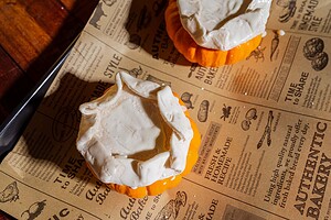 Two small pumpkins with cream-colored pastry dough draped over the tops, resembling puff pastry tops, are placed on a vintage-style printed baking sheet—perfect for serving pumpkin soup in charming mini pumpkin bowls.