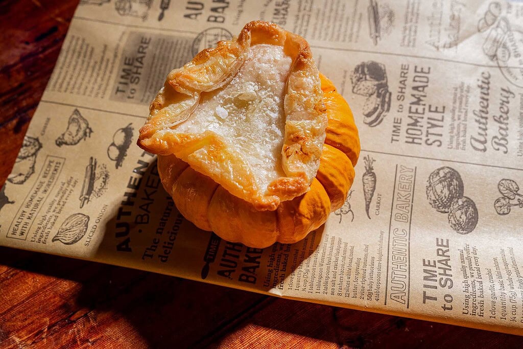 A small pumpkin filled with a baked pastry crust sits on a sheet of brown paper with printed bakery-themed text and illustrations, resembling one of those charming mini pumpkin bowls topped with golden puff pastry tops—the perfect vessel for serving warm pumpkin soup.