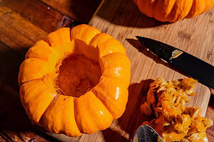 A small pumpkin with the top cut off sits on a wooden board next to pumpkin seeds, pulp, a knife, and a spoon—perfect for making charming mini pumpkin bowls for serving creamy pumpkin soup.