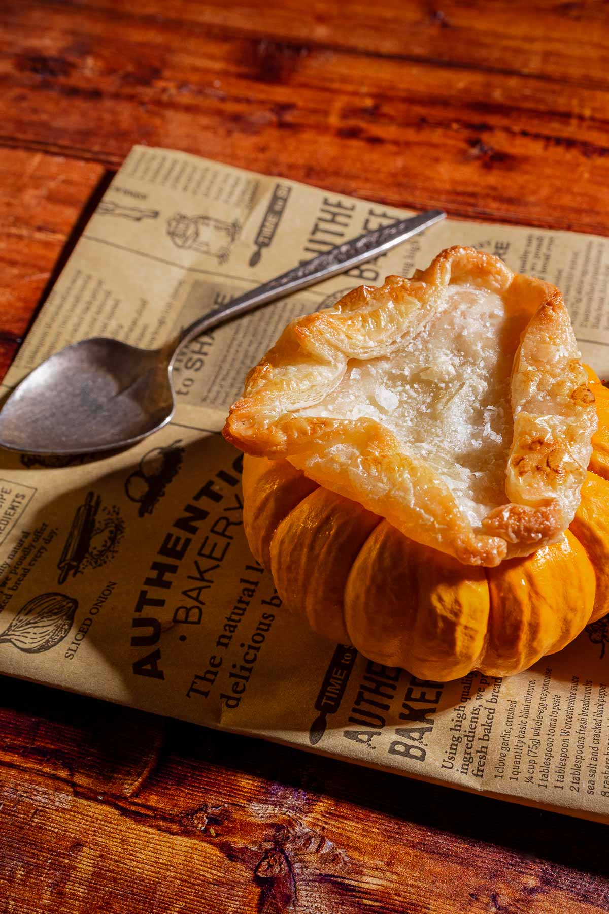 A baked pastry with a golden crust, crafted from delicate puff pastry tops, sits atop a small pumpkin, creating charming mini pumpkin bowls placed on printed parchment paper next to a metal spoon on a wooden surface.