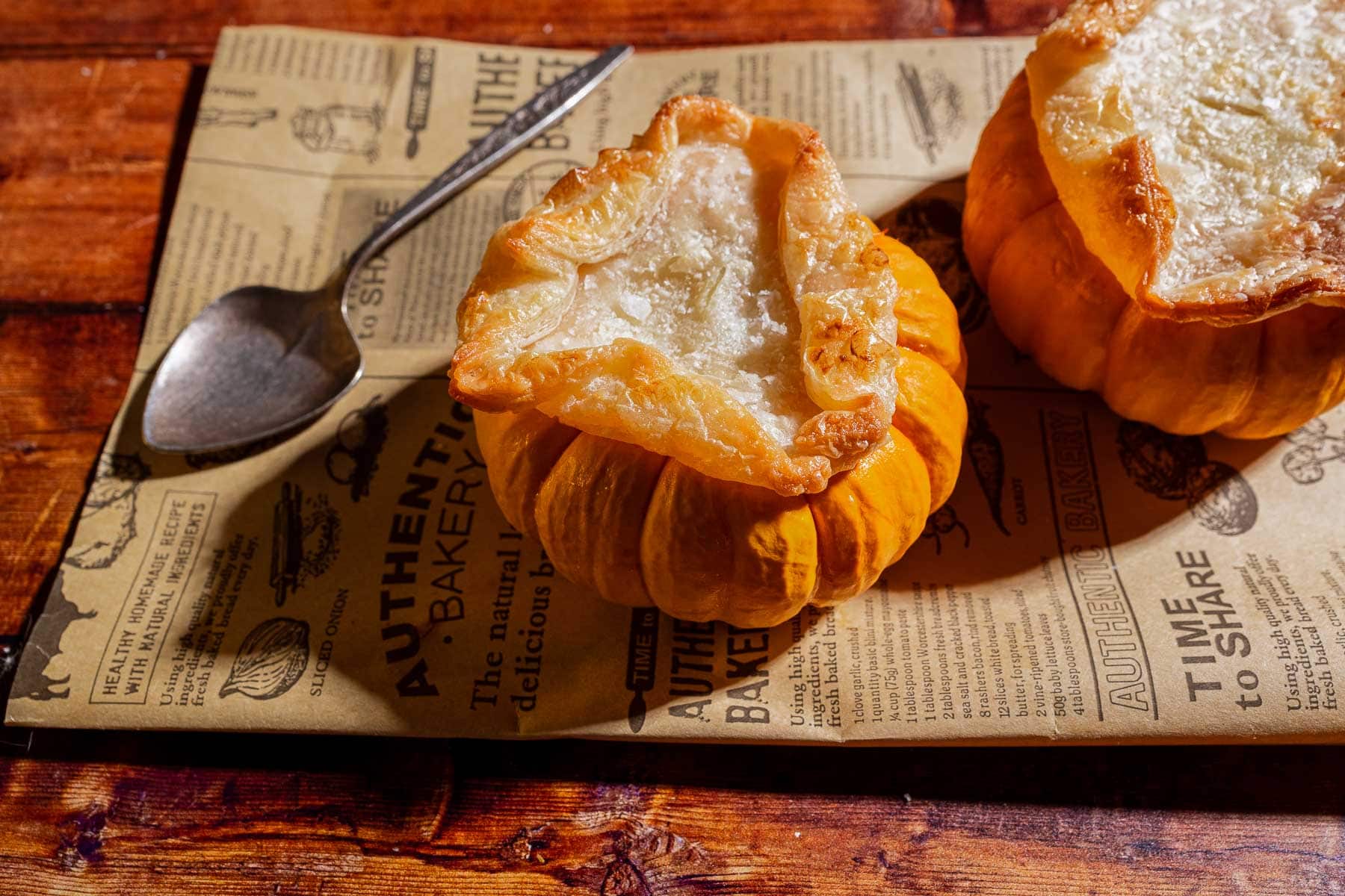 Two mini pumpkin bowls filled with rich pumpkin soup and topped with golden puff pastry tops are placed on printed parchment paper beside a metal spoon.
