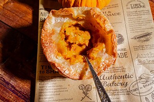A spoon scoops out creamy pumpkin soup from a round mini pumpkin bowl placed on bakery-themed parchment paper.