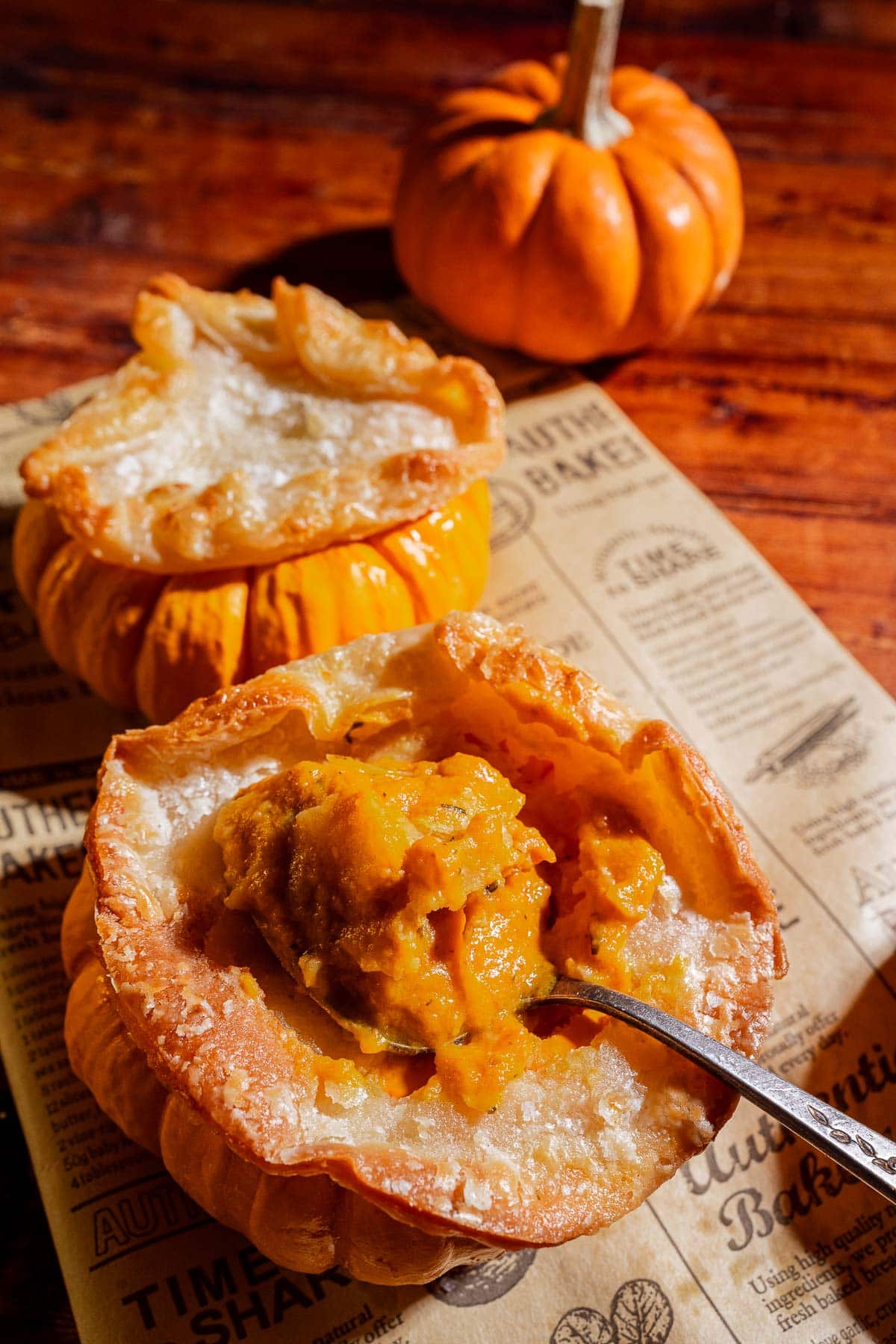 A baked mini pumpkin bowl filled with creamy pumpkin soup and topped with a golden puff pastry top, served on a sheet of printed parchment paper with another whole mini pumpkin in the background.