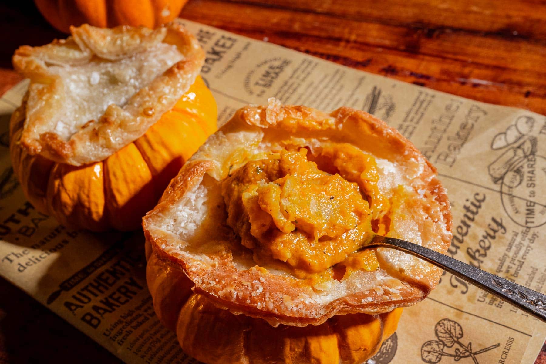 Mini pumpkin bowls filled with pumpkin soup and topped with flaky puff pastry tops, one with a spoon scooping out the soup, set on bakery-themed paper.
