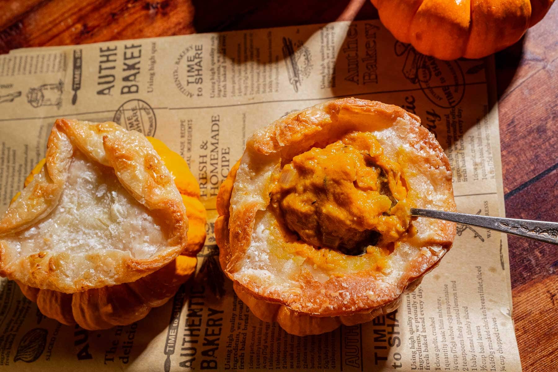 Two small mini pumpkin bowls with their tops removed are filled with a baked, creamy orange pumpkin soup; one is being scooped with a spoon, both set on vintage-style parchment paper.