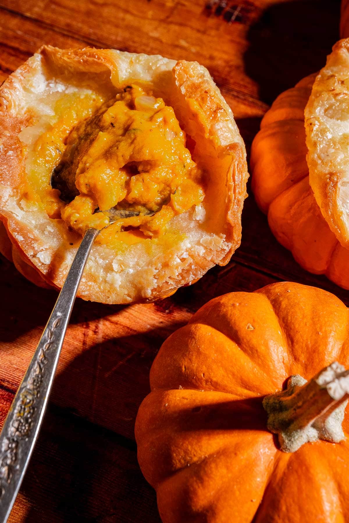 A spoon scoops pumpkin soup from a baked pastry shell with puff pastry tops, while two small whole pumpkins—perfect for mini pumpkin bowls—sit nearby on a wooden surface.