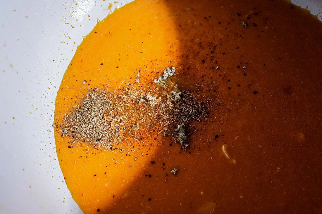 Orange liquid with piles of ground spices and minced garlic in a white bowl, partially covered by a shadow—perfectly reminiscent of creamy pumpkin soup, ready to be served in charming mini pumpkin bowls or topped with golden puff pastry tops.