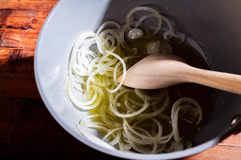 Thinly sliced onions in a gray bowl with olive oil and a hint of apple chili crisp, being stirred with a wooden spoon on a wooden surface.