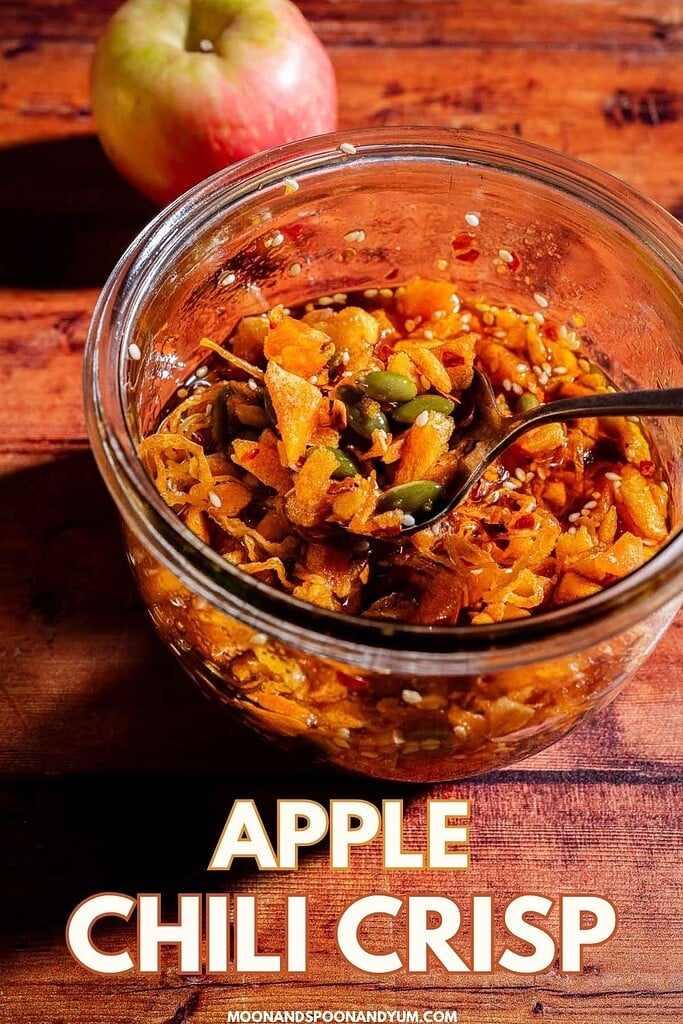 A glass jar of apple chili crisp with a spoon sits on a wooden surface; an apple is in the background, highlighting the sweet and spicy allure of this unique apple chili crisp spread.