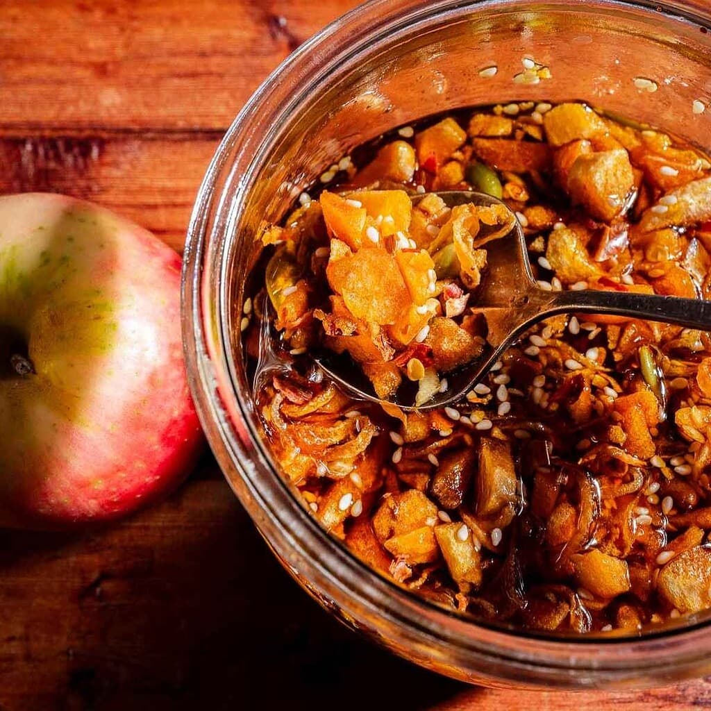 A glass jar of chunky apple chili crisp chutney with visible sesame seeds, next to a whole apple on a wooden surface.