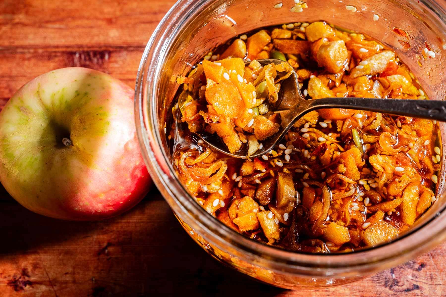 A spoon lifts chunky apple and ginger pieces from a jar of homemade apple chili crisp condiment with sesame seeds, next to a whole apple on a wooden surface.
