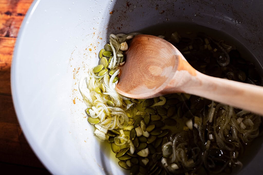 A wooden spoon rests in a white bowl containing sliced shallots, pepitas, chopped garlic, olive oil, and a dollop of apple chili crisp.